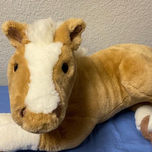 Puede incluir: Un juguete de peluche de caballo marrón y blanco acostado sobre una manta azul. El caballo tiene la cara blanca y una crin blanca.