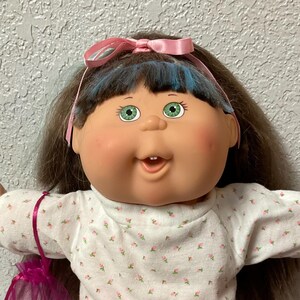 May include: A doll with brown hair, green eyes, and rosy cheeks, wearing a white long-sleeved dress with a small pink floral pattern and a ruffled hem. The doll has a pink ribbon in its hair, white shoes with lace trim, and a small purple organza bag.