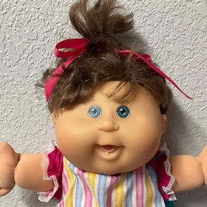 May include: A vintage Cabbage Patch Kid doll with brown hair styled in a high ponytail, adorned with a pink ribbon. The doll wears a striped top and pants in pink, yellow, and blue, paired with pink shoes and lace-trimmed socks.
