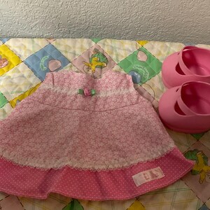 May include: A sleeveless pink dress featuring a white floral pattern, a white lace trim across the chest, a small green bow, and a polka-dot ruffle hem, accompanied by a pair of solid pink plastic shoes with a single strap across the top.