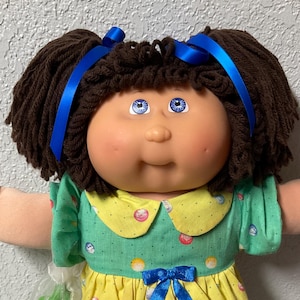 May include: A vintage Cabbage Patch Kid doll with brown yarn hair tied with blue ribbons. The doll wears a green and yellow dress with a blue bow, white lace trim, and white shoes. The doll has blue eyes and rosy cheeks.