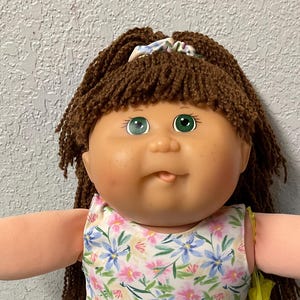 May include: A vintage doll with brown yarn hair, green eyes, and freckles. The doll wears a floral print dress with pink, blue, and white flowers. It has light green socks and white shoes. A yellow accessory is visible on the back.