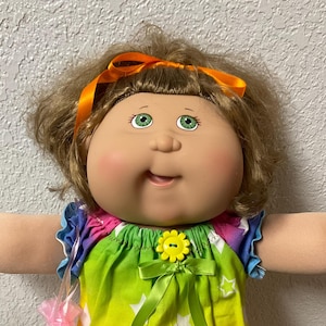 May include: A doll with light brown hair, green eyes, and rosy cheeks, wearing an orange ribbon in its hair. The doll is dressed in a rainbow-colored top with a white star pattern, a yellow flower button, a green ribbon bow, and white lace-trimmed pants.