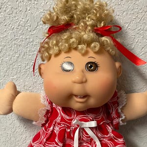 Op de afbeelding: Een pop met blond krullend haar vastgebonden met rode linten, met één zilveren oog en één bruin oog. De pop draagt een rode jurk met een wit cirkelpatroon, een wit lintstrikje bij de halslijn en witte kanten randen aan de mouwen en zoom.
