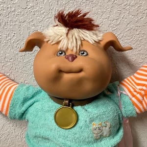 May include: A vintage doll with a brown face, floppy ears, and a tuft of brown and white hair. It wears a teal shirt with a cat patch, orange and white striped pants and matching arm warmers. A gold medallion hangs from its brown collar.
