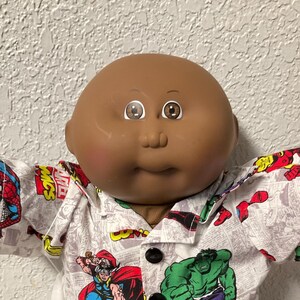 May include: A Cabbage Patch Kid doll wearing a button-down shirt with Marvel comic book characters and dark blue denim shorts. The doll has brown skin, brown eyes, and white sneakers.