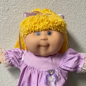 May include: A doll with bright yellow yarn hair tied with a purple ribbon, featuring blue eyes and a round face. The doll wears a light purple dress with white lace trim on the sleeves and front, white ruffled socks, and white shoes.