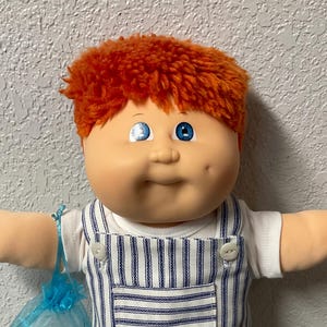 May include: A vintage doll with orange hair and blue eyes, wearing a white shirt and blue and white striped overalls. The doll has a soft, plush body and is holding a small blue bag.