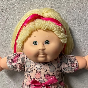 May include: A Cabbage Patch Kid doll with blonde yarn hair and a pink ribbon. The doll wears a pink dress with a cat pattern and a matching pink bow. White shoes and socks complete the outfit. The doll is on a colorful quilt.