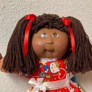 May include: A vintage Cabbage Patch Kids doll with brown yarn hair tied with red ribbons. The doll wears a red dress with a white bow and a Hello Kitty print. The doll has brown skin and is wearing white socks and shoes.