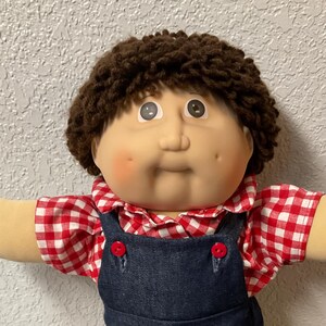 May include: A vintage doll with a soft, round face, brown curly hair, and rosy cheeks. The doll wears a red and white checkered shirt under dark blue denim overalls with red buttons. White sneakers complete the outfit.