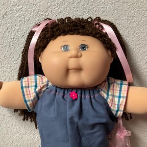 以下が含まれることがあります： 茶色の巻き毛、青い目、丸い顔をしたヴィンテージ人形。 人形は、チェック柄のトリムとピンクの花のアクセントが付いた青いデニムのドレスを着ています。 ピンクのリボンが髪を飾り、人形はピンクのフリルソックスと白い靴を履いています。