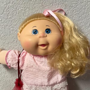 Peut inclure: Une poupée Cabbage Patch Kids vintage avec des cheveux blonds et des yeux bleus. La poupée porte une tenue rose avec une bordure en dentelle, un nœud rose et des chaussures roses. Un sac en filet rouge est suspendu au bras de la poupée.