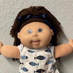 May include: A vintage doll with brown hair and blue eyes, wearing a white dress patterned with blue and black fish. The doll has a blue ribbon in its hair and white shoes. A small white bag hangs from its arm.