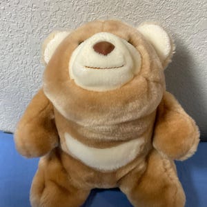 Könnte beinhalten: Ein hellbrauner Teddybär mit einem weißen Bauchfleck und einer braunen Nase. Der Bär hat kleine, abgerundete Ohren und ein gesticktes Lächeln. Er sitzt auf einer blauen Oberfläche.
