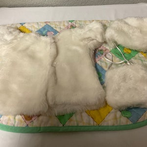May include: A white faux fur baby doll outfit with a jacket, hat, and a small purse. The outfit is laid out on a green and yellow quilted blanket.