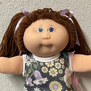 May include: A vintage doll with brown yarn hair styled in pigtails, adorned with purple ribbons. The doll wears a floral print dress in shades of gray, yellow, and white, paired with white lace socks and shoes. The doll has blue eyes.