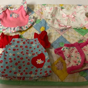 Puede incluir: Cuatro conjuntos de ropa para una muñeca bebé. Los conjuntos son de color rosa, azul y blanco con estampados florales y de fresas. Los conjuntos están sobre una colcha verde y blanca.