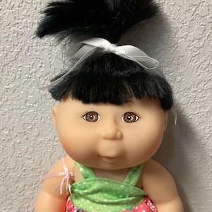 May include: A vintage doll with black hair styled in a high ponytail, secured with a white bow. The doll wears a pink floral dress with a green top and white lace trim. It has brown eyes and is holding a small, clear bag.