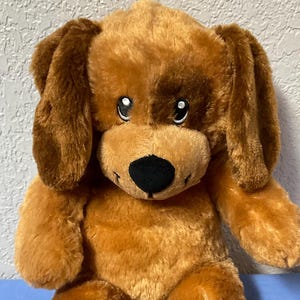 May include: A brown plush dog with large, floppy ears, a black nose, and white eye accents. The soft toy is positioned upright on a blue surface. The dog has big, soft paws and a fluffy body.