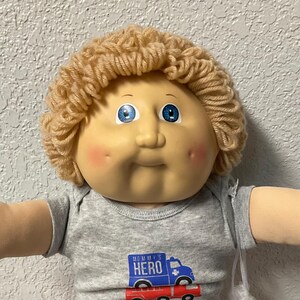 Könnte beinhalten: Eine Cabbage Patch Kid-Puppe mit blonden Garnhaaren, blauen Augen und rosigen Wangen. Die Puppe trägt einen grauen Strampler mit einem "Mommy's Hero"-Aufdruck, eine rote Hose und weiße Schuhe. Eine transparente Plastiktüte ist befestigt.