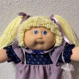 May include: A vintage doll with blonde yarn hair styled in pigtails, wearing a lavender dress with a navy blue floral bodice. The doll has blue eyes and is wearing matching lavender socks and shoes with lace trim. A small bag is attached.