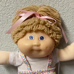 May include: A doll with light skin, blue eyes, and dimples, featuring blonde yarn hair tied with a pink ribbon. The doll wears a white short-sleeved shirt under a white jumper with a colorful heart pattern, paired with pink ruffled socks and white shoes.