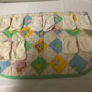 Może przedstawiać: Osiem par białych skarpetek dla niemowląt na zielono-białym pikowanym kocyku z kolorowym wzorem kreskówkowym przedstawiającym dzieci i balony.