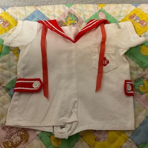 May include: White sailor-style romper with red detailing. Includes a red collar, red ribbon, and red trim on the shorts. A small pocket with a red anchor emblem is on the chest. Buttons are visible on the shorts.