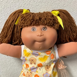 May include: A doll with brown yarn hair styled in two pigtails with yellow ribbons. The doll wears a sleeveless yellow top featuring a pattern of bears, yellow ducks, and purple flowers, paired with white lace-trimmed socks and purple shoes.