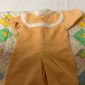 Op de afbeelding: Een licht oranje baby romper van corduroy met een witte gehaakte kraag.