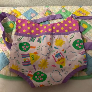 May include: A pink and white baby bib with a purple trim and a colorful pattern of suns, clouds, trees, and flowers. The bib has the letters "CPK" printed on it.