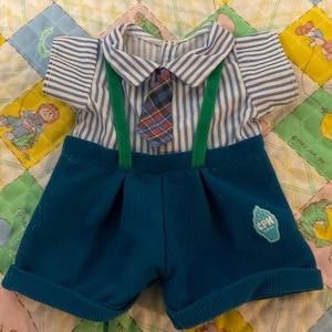 May include: A doll's outfit comprising a blue and white striped shirt with a plaid tie and green braces. The outfit includes teal shorts with a logo patch. The outfit is displayed on a colourful quilt.