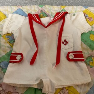 May include: White sailor-style romper with red detailing. Includes a red-trimmed collar, red ribbon ties, and red trim on the shorts. An embroidered red anchor is on the pocket. White buttons with a red border. The romper is on a colourful quilt.