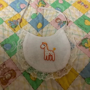 May include: White baby bib with a lace trim and an embroidered giraffe design. The bib is on a quilted fabric with a checkered pattern and colourful cartoon characters.