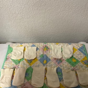 Może przedstawiać: Kolekcja kremowych skarpetek dla niemowląt ułożonych na kolorowej, pikowanej macie do zabawy. Mata ma wzór patchworkowy z pastelowymi kwadratami i zieloną obwódką. Skarpetki są starannie ułożone, co sugeruje artykuł dla dziecka.