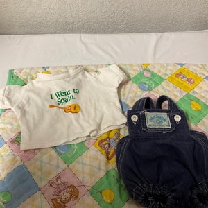 Könnte beinhalten: Ein weißes T-Shirt mit dem Text "I Went to Spain." und einer Gitarrengrafik. Eine blaue Jeanslatzhose mit einem Patch, auf dem "Care Bears." steht. Die Artikel liegen auf einer grün-gelben Patchworkdecke.