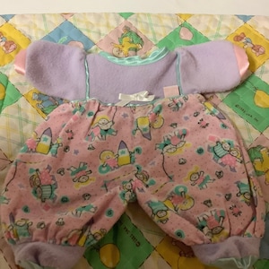 Puede incluir: Un conjunto de muñeca bebé rosa y morado con una camisa de manga larga y pantalones. Los pantalones tienen un estampado de niñas pequeñas jugando con una tabla de surf. La camisa tiene un ribete azul claro y un lazo blanco.