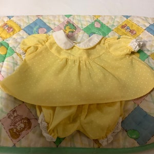 May include: A yellow baby outfit with white polka dots, comprising a collared top and matching bloomers. The outfit has white lace trim and is placed on a colourful patchwork quilt.