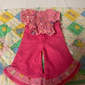 May include: Pink and white gingham doll's clothes with a frilled hem. The top has a floral print and the trousers have a frilled hem.