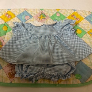 May include: A blue and white gingham dress and bloomers set for a doll. The dress has a white collar and short sleeves. The bloomers have a ruffled hem.