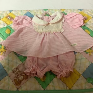 May include: A pink and white striped dress and bloomers set for a doll. The dress has a white collar and lace trim. The bloomers have a ruffled hem.