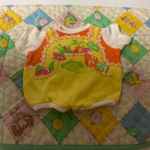 May include: A yellow and orange baby onesie with a white trim. The onesie features a picture of vegetables and a garden scene. The text "Cabbage Patch Kids" is printed on the onesie.