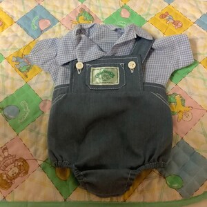 May include: A blue and white checked shirt and blue denim dungarees with a green label that says "Cabbage Patch Kids" on it. The outfit is laid out on a colourful quilt.