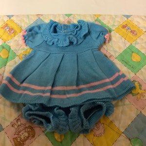 May include: A blue knitted baby dress with pink trim and a ruffled collar. The dress has a matching blue knitted nappy cover.