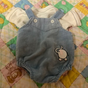 Op de afbeelding: Babyblauwe corduroy romper met witte mouwen en een olifantenapplicatie. De romper heeft twee witte knopen en ligt op een kleurrijke patchwork quilt.