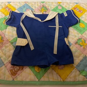 May include: A blue sailor-style baby onesie with white trim and white ribbon ties. The onesie has a white button on the front and a white pocket on the side.