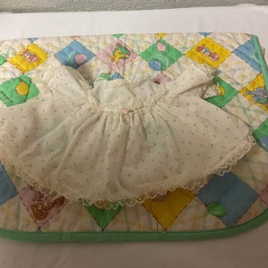 May include: A white baby doll dress with a lace trim and a small pink flower accent. The dress is on a colourful quilted blanket with a checkered pattern.