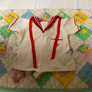 May include: White sailor-style onesie with red trim and red ribbon ties. The onesie has a small pocket with a red anchor design.