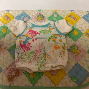 May include: A white Cabbage Patch Kids onesie with a blue paintbrush and colourful paint splatters. The onesie has the words "Cabbage Patch Kids" and "Made in USA" printed on it.